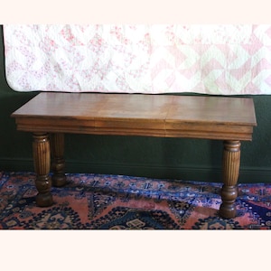 May include: A rectangular wooden table with a light brown finish. The table has four decorative, fluted legs. A pink and white quilt hangs in the background. The table is approximately 152 cm long.