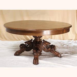 May include: A round wooden table with a smooth, circular top and a central pedestal base. The base features intricate carvings of three lion heads, each extending into a carved leg that terminates in a paw-shaped foot, finished in a rich brown.