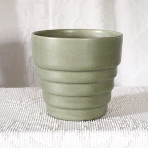 May include: A sage green ceramic planter with a stepped, ridged design. The cylindrical pot has a wide opening and is suitable for indoor or outdoor use. The neutral color complements various decor styles. A decorative item for plants.