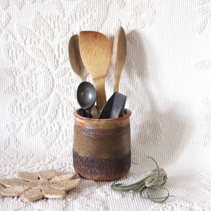 Puede incluir: Un porta utensilios de cerámica marrón con un diseño rústico, que contiene cucharas de madera y un cucharón de metal. El porta utensilios está sobre una superficie blanca con una estera tejida.