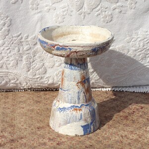 May include: A white ceramic bird bath with blue and brown painted accents. The bath is on a pedestal with a wide base.