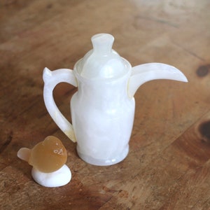 May include: A white ceramic teapot with a lid and a handle. The teapot is sitting on a wooden table next to a small, brown bird figurine.