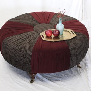 May include: A round ottoman with a patterned brown and burgundy fabric design. A small brass tray with two red apples and a small vase with a few sprigs sits on top. The ottoman has small, decorative legs.