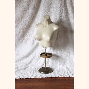 Könnte beinhalten: Eine weiße Gips-Torso-Schaufensterpuppe auf einem goldenen Metallständer. Der Schaufensterpuppe fehlen Arme und Kopf. Der Ständer hat eine runde Basis und ein rundes Regal.
