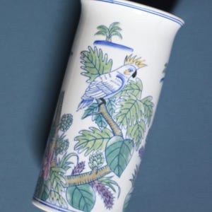 Könnte beinhalten: Weiße Keramikvase mit einem blau-grünen Papageien- und tropischen Blattmuster. Die Vase hat einen blauen Rand und Boden mit einer detaillierten Illustration eines Papageis auf einem Ast.
