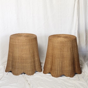 May include: Two matching woven wicker side tables. The tables have a round top and a flared, scalloped base. The wicker is a light brown color, and the tables are set against a white background.