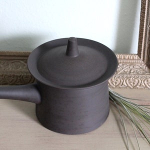 Puede incluir: Una tetera de cerámica marrón oscuro con tapa y asa. La tetera está sobre una superficie de madera marrón claro.