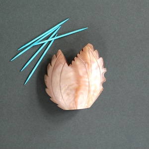 May include: A light brown, leaf-shaped dish with a textured surface. Four blue toothpicks are placed on a dark gray surface next to the dish.