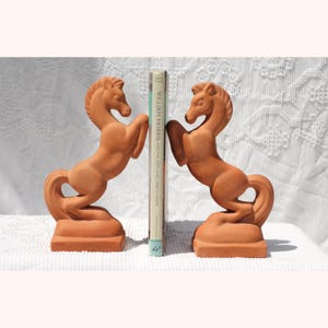 May include: A pair of terracotta horse bookends. Each horse is rearing up on its hind legs, with a curved tail and a detailed mane. The bookends are flanking a book with the title "WILLIAM MORRIS AND THE ARTS AND CRAFTS HOME".