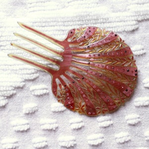 Puede incluir: Un peine para el cabello vintage con un diseño de concha. El peine está hecho de plástico y tiene un color marrón rojizo con detalles dorados. El peine tiene tres dientes y está decorado con pequeños puntos negros.