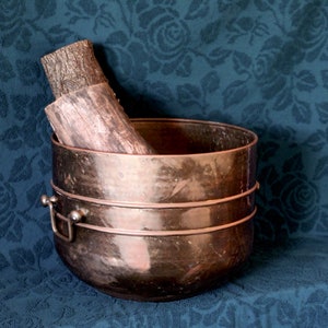 May include: A copper fire basket with two pieces of firewood inside. The basket has two metal bands around the top and two handles on the sides. The basket is sitting on a blue fabric with a floral pattern.