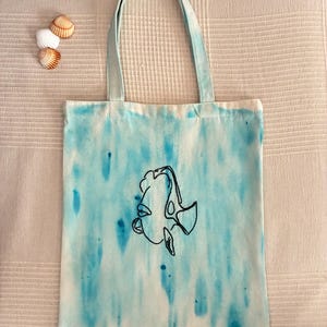 May include: A white tote bag featuring a blue watercolor-style tie-dye pattern and a black line-art illustration of a fish in the center. The bag has two matching blue fabric handles and is accompanied by three small seashells placed to the left.