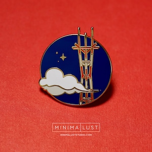 May include: Enamel pin featuring a blue circle with a gold border, depicting a stylized tower with red and gold accents, a white cloud, and a gold star. The pin has the text "MINIMA LUST" and "minimaluststudio.com" in white.