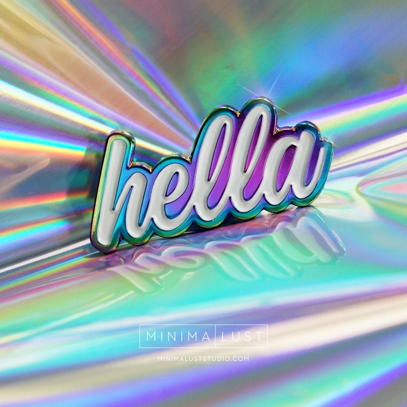 May include: A shiny enamel pin with the word "hella" in white, outlined in iridescent colors and a gold border. The pin is set against a holographic background, reflecting various colors. The pin is a decorative accessory.