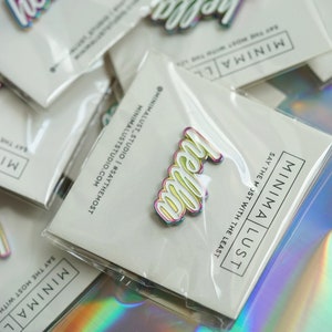 May include: A collection of iridescent "hello" pins with white outlines, packaged on cards with the "MINIMA LUST" logo and the text "SAY THE MOST WITH THE LEAST". The pins have a rainbow color effect.