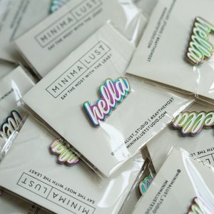 May include: A collection of iridescent "hella" enamel pins on white backing cards. The pins have a white outline and a rainbow gradient effect. The cards are packaged in clear plastic and feature the "MINIMA LUST" logo and social media handles.