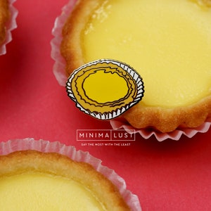 May include: A close-up shot of a yellow enamel pin shaped like an egg tart, placed on a red surface near two egg tarts. The pin has a yellow center with a black outline and a white and black scalloped edge. The text "MINIMA LUST" and "SAY THE MOST WITH THE LEAST" is visible.