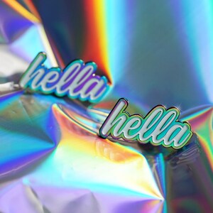 May include: Two enamel pins with the word "hella" in a cursive font. The pins have a white base with a holographic, iridescent outline and text. The background is a reflective, rainbow-colored surface.