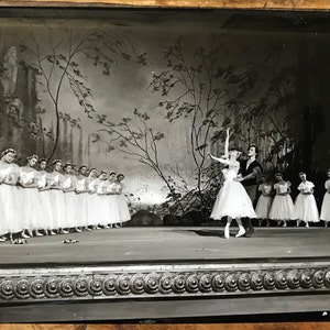 Może przedstawiać: Czarno-białe zdjęcie przedstawiające przedstawienie baletowe. Tancerka w białym tutu jest unoszona przez tancerza. Grupa tancerek w białych tutu stoi w rzędzie w tle.