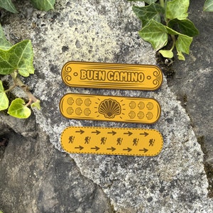 May include: Three yellow decorative stickers with a Spanish phrase, a seashell design, and arrows. The stickers say "BUEN CAMINO" which means "good journey" in Spanish.