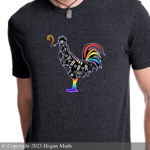 Op de afbeelding: Een zwart T-shirt met een kleurrijk haanontwerp. De haan houdt een sigaret in zijn snavel en heeft een regenboogstaart.