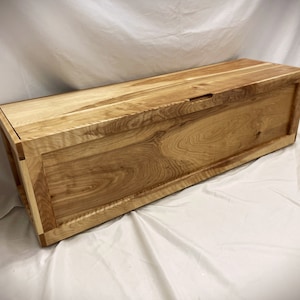 May include: A wooden storage chest with a natural wood finish and a hinged lid. The chest has a simple design with a rectangular shape and a subtle grain pattern.