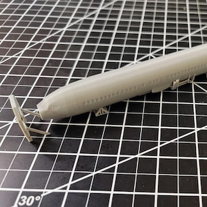 Könnte beinhalten: Ein graues Plastikmodell eines Flugzeugs mit abnehmbarem Heck. Das Modell befindet sich auf einer schwarz-weißen Gitteroberfläche.