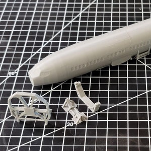 May include: A gray plastic model airplane kit with multiple parts, including the fuselage, wings, tail, and landing gear. The kit is laid out on a black and white grid surface.