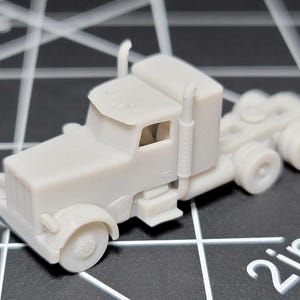 May include: A small, light grey model of an articulated lorry. The truck features a cab, a trailer hitch, and dual rear wheels. The model is approximately 5 cm long.