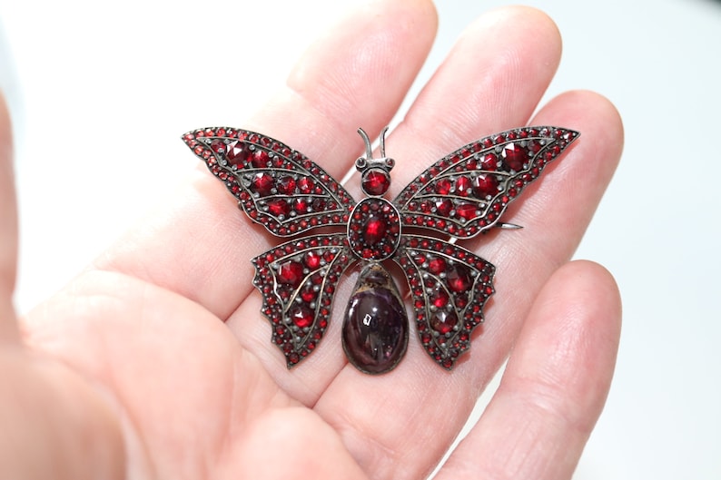 May include: A silver butterfly brooch with red garnet stones. The butterfly has a large, teardrop-shaped garnet in the center.