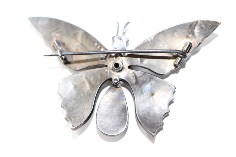 May include: Silver butterfly brooch with a pin on the back. The butterfly has a detailed design and a teardrop-shaped body.