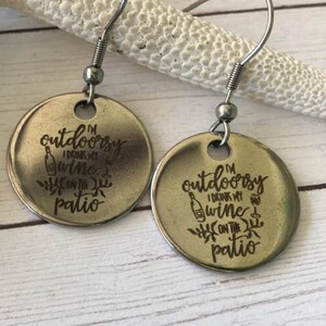 May include: A pair of silver-toned, round earrings with a hook closure. Each earring features the engraved text "I'm outdoorsy, I drink my wine on the patio" with a wine bottle and floral design. The earrings are displayed on a white starfish.