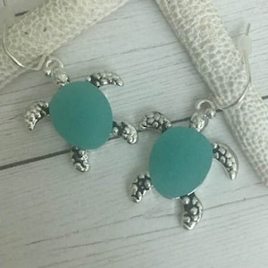 May include: A pair of silver turtle earrings with teal blue glass accents. The earrings are hanging from a white starfish on a white wooden background.