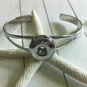 May include: A silver-colored cuff bracelet with a large, round, silver snap closure. The bracelet is resting on a white starfish against a light wood-grain background. The bracelet has a simple, modern design.