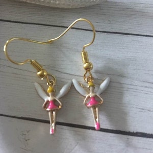May include: A pair of gold-toned hook earrings featuring small fairy charms. Each charm has white wings, a bright pink dress, and a yellow head. The earrings hang from gold-colored metal hooks, showcasing a detailed design with vibrant enamel accents.
