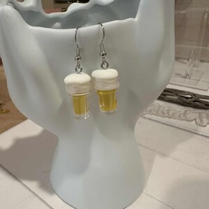 May include: A pair of novelty earrings shaped like miniature beer glasses. Each earring features a clear, yellow-tinted glass with a white foam top, suspended from a silver-colored fish hook. The earrings are displayed against a white background.