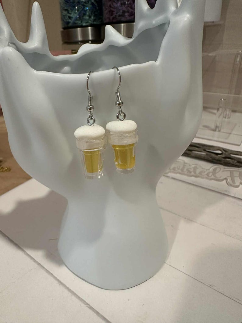 May include: A pair of novelty earrings shaped like miniature beer glasses. Each earring features a clear, yellow-tinted glass with a white foam top, suspended from a silver-colored fish hook. The earrings are displayed against a white background.