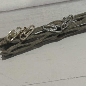 May include: Two pairs of paperclip-shaped stud earrings. One pair is gold-toned and the other is silver-toned. Both pairs are encrusted with small, clear crystals. The earrings are displayed on a piece of driftwood against a white background.