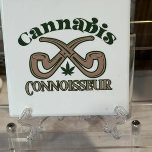May include: White square tile with the word "cannabis" in green script above two crossed brown pipes and a green cannabis leaf. The word "CONNOISSEUR" is printed below the pipes in green.