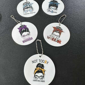 May include: White round keychains with illustrations of women with glasses and headbands. Each keychain has a different design, including text that reads "Diabetes Warrior", "Believe", "Epilepsy Warrior", "Multiple Sclerosis Warrior", and "Not Today Endometriosis Warrior".