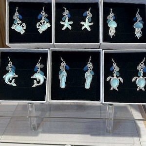 May include: A display of silver-toned earrings with ocean-themed charms, including crabs, starfish, jellyfish, dolphins, flip-flops, and turtles. Each earring set is presented in a small white box. The earrings have blue and clear bead accents. The text "@Twistedflamingo_954" is visible.