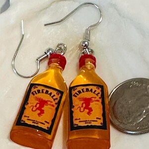 Puede incluir: Un par de pendientes novedosos con dijes en miniatura de botellas de whisky Fireball. Los pendientes tienen ganchos para las orejas plateados y tapas rojas. Las botellas tienen un l&iacute;quido amarillo-naranja y una etiqueta con la palabra "Fireball".
