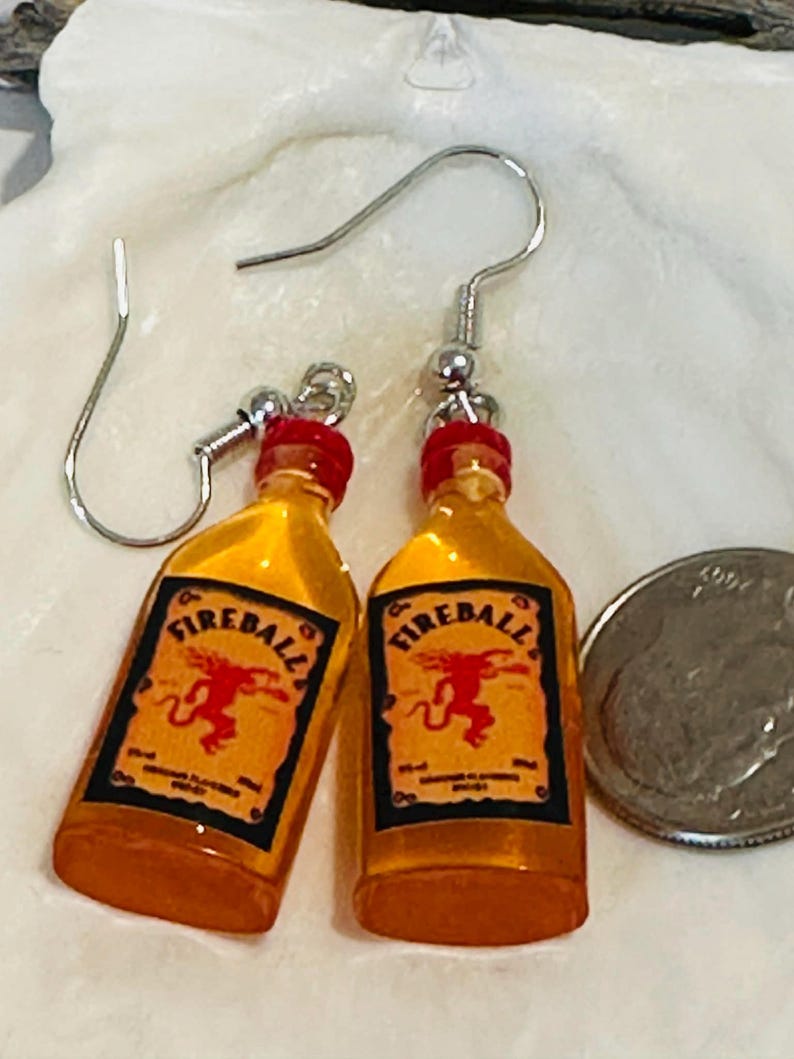 Puede incluir: Un par de pendientes novedosos con dijes en miniatura de botellas de whisky Fireball. Los pendientes tienen ganchos para las orejas plateados y tapas rojas. Las botellas tienen un l&iacute;quido amarillo-naranja y una etiqueta con la palabra "Fireball".