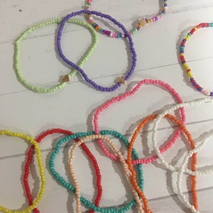 May include: A collection of colorful beaded bracelets with gold heart charms. The bracelets are made with small, round beads in various colors, including green, purple, pink, orange, yellow, turquoise, and white.