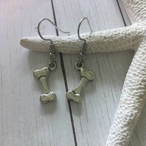 May include: A pair of silver-colored dog bone earrings. Each earring features a bone-shaped charm suspended from a simple hook. The earrings are displayed on a white starfish and a light wood-grain surface.