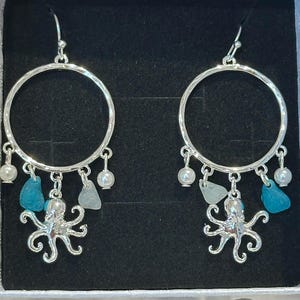 May include: Silver hoop earrings with an octopus charm, sea glass, and small white pearls. The earrings are displayed in a white box with a black interior. The hoops have a hammered texture.