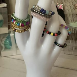 May include: A collection of beaded rings in various colors, including gold, green, pink, blue, and iridescent. The rings are displayed on a white hand-shaped jewelry holder, showcasing their different designs and sizes. The rings are a mix of colorful and neutral tones.
