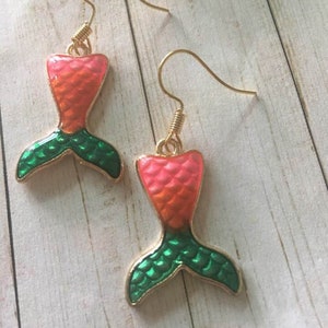 May include: A pair of gold-toned earrings featuring mermaid tail charms. The charms are in the shape of a mermaid tail, with a gradient of pink to orange, and green tail fins. The earrings are on a light wood grain surface.