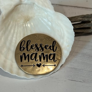 May include: A round, gold-colored metal disc with the words "Blessed Mama" in black script, a heart, and arrows. The disc rests inside a white seashell, with a wooden element in the background.