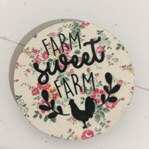 May include: A round wooden sign with a floral pattern and the text "Farm Sweet Farm" in black lettering. A black silhouette of a rooster is in the center of the sign.